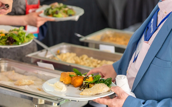 A close-up of a person serving food onto a plate at a buffet, with the plate in clear focus.