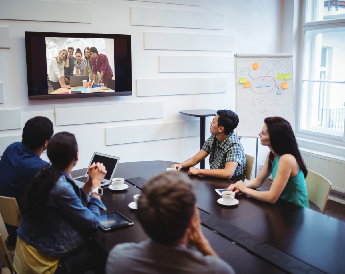 Group of people sitting at a table, looking at a tv on the wall showing another group of people