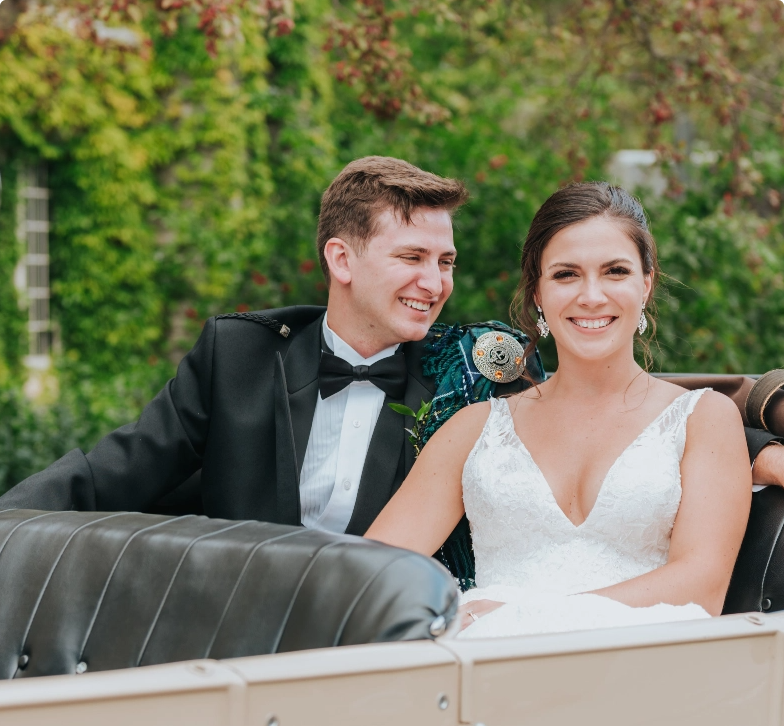 Two people dressed in wedding attire, sitting in a vintage car.