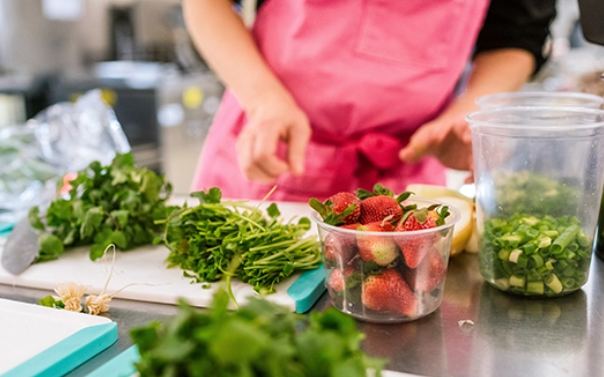 Person in a pink apron preparing fresh ingredients, including strawberries, herbs and green onions, on a kitchen countertop.